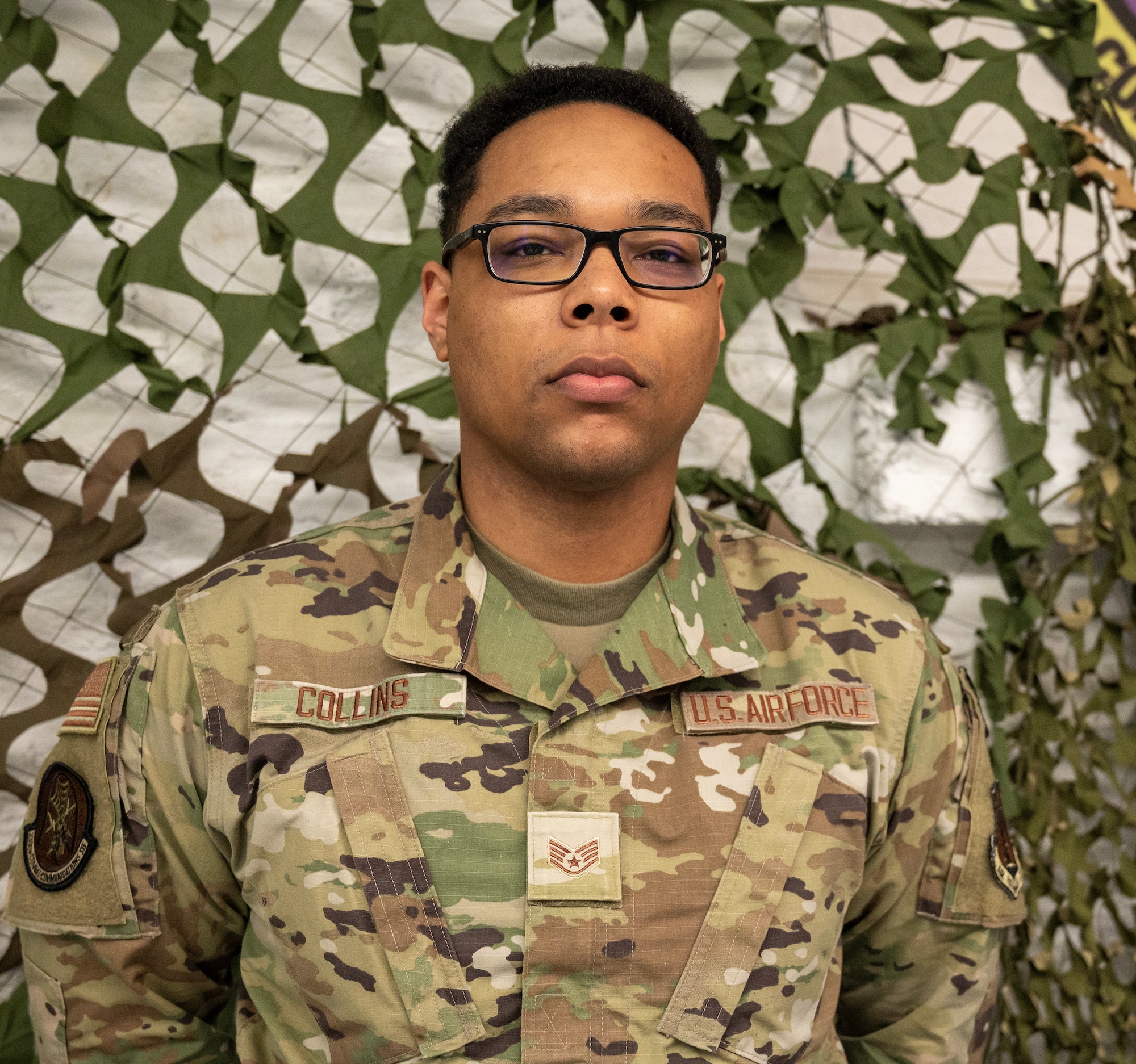 A U.S. Air Force Airman poses for a photo in front of a camoflague web netting background. He is wearing an OCP pattern uniform.