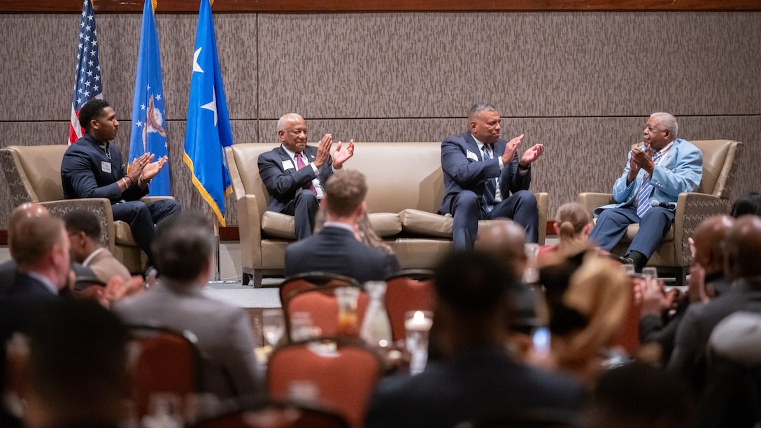 Four people clap on stage with a crowd of others in the audience during the Accelerating the Legacy event at Joint Base Charleston, South Carolina, Feb. 16, 2024.