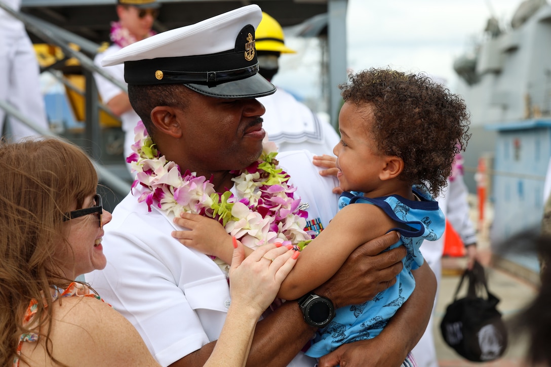 A sailor smiles while holding a child. A second person stands and smiles on the sailor's left.