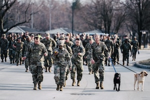 Maj. Gen. Martin walks in a row with three other Airmen and his two leashed dogs, a larger group of Airmen follow behind.