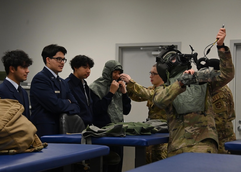Man wearing helmet and gas gear protection equipment shows how to use it to a group of high school students.