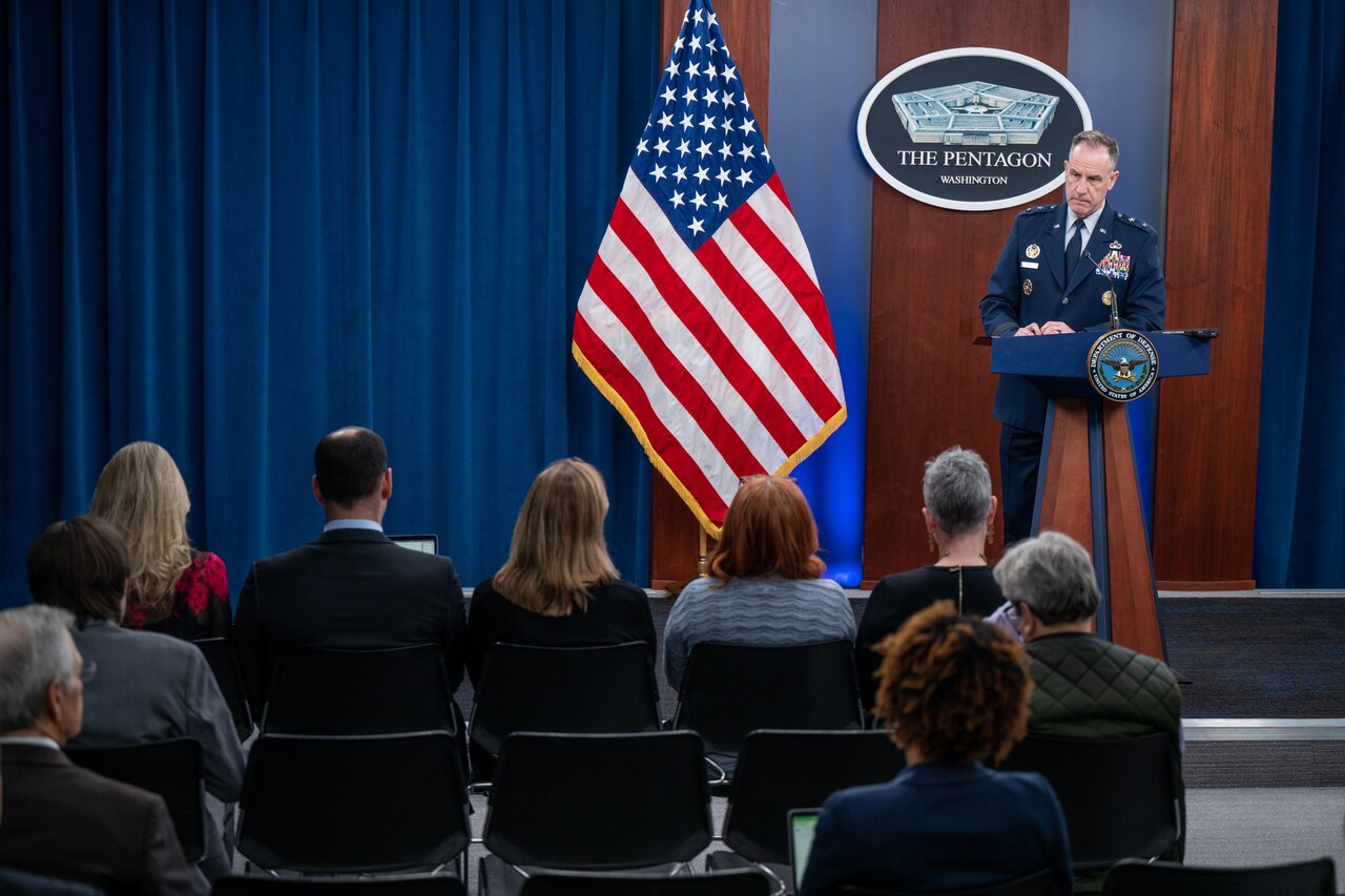 A man in a military uniform stands behind a lectern and addresses a seated audience of reporters.