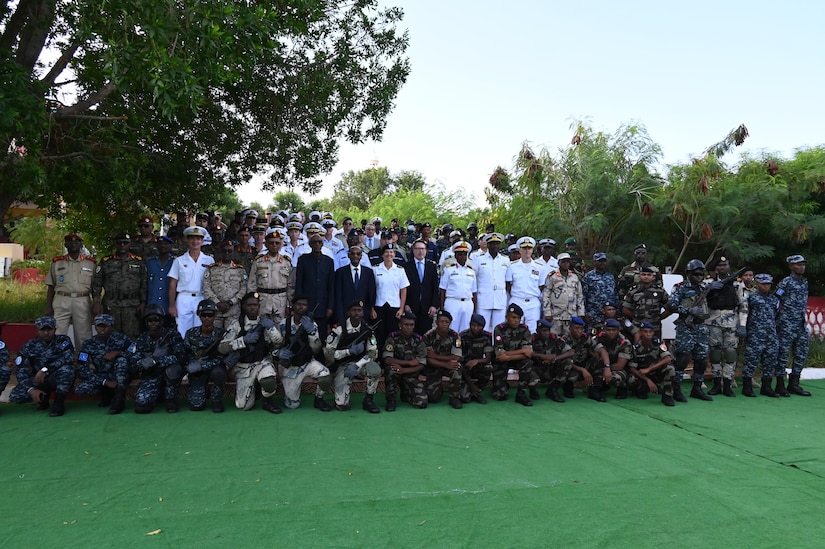 A group of military members and civilians pose for a photo on a green with trees in the background.