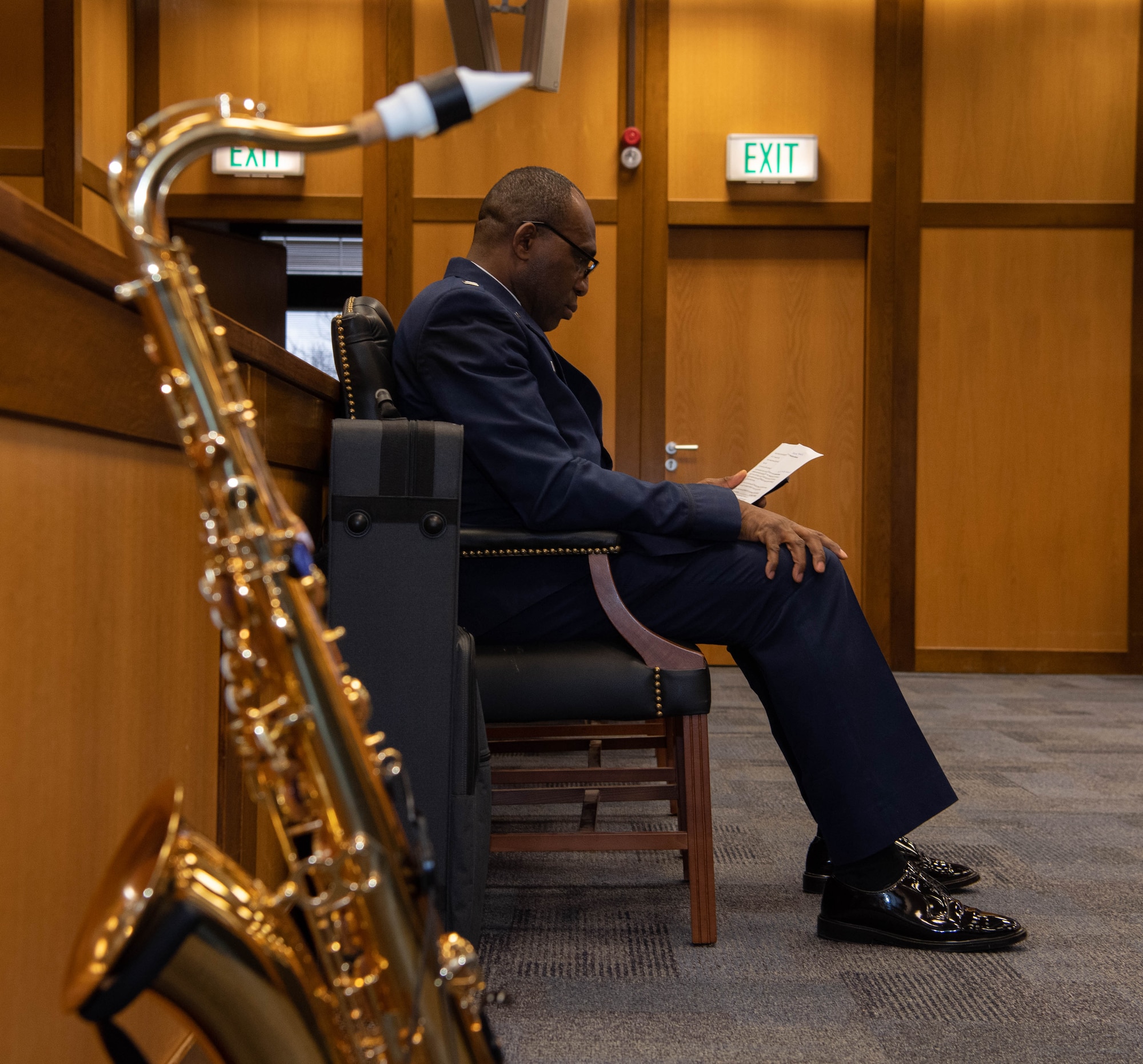 Man reads a paper in the background, a saxophone is shown in the foreground