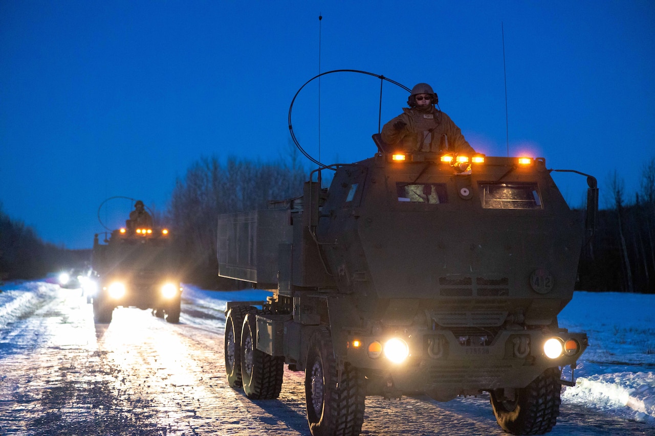 Marines stand in large military vehicles travelling down a snow-covered road with headlights on at night.