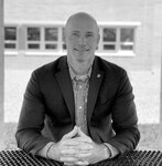 A black and white photo of a light skinned man seated at a outdoor table. He is bald.