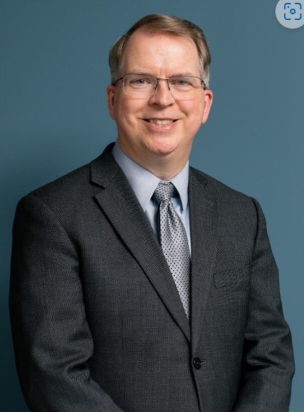 A light skinned man with graying hair and glasses in a grey suit.