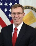 A light skinned man with brown hair and glasses in a dark suit.