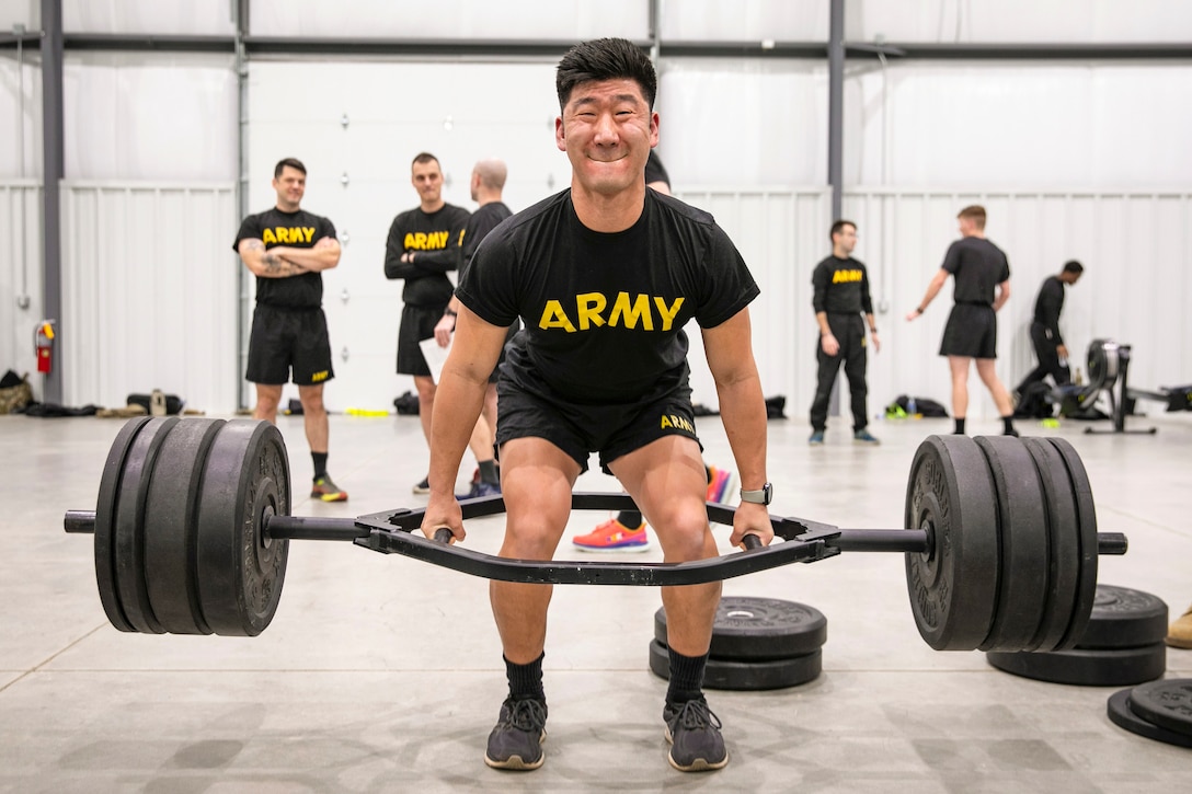 A soldier conducts a deadlift with heavy weights as fellow soldiers watch in the background.