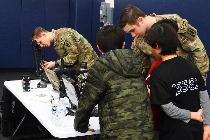 Two soldiers standing behind a table work with electronics while two children lean in to observe.