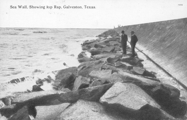 A vintage postcard of the Galveston Sea Wall shows two men standing on rip rap, which is rock or other material used to protect shoreline structures against scour and water, wave, or ice erosion.