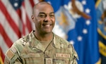 Head and shoulders image of a military person with flags in the background blurred.