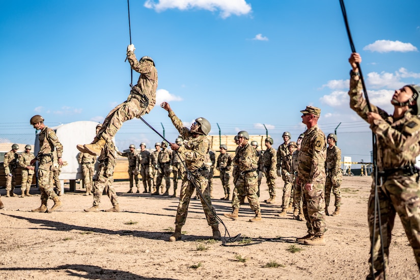 A soldier helps a fellow soldier rappel down a tower as fellow soldiers watch.