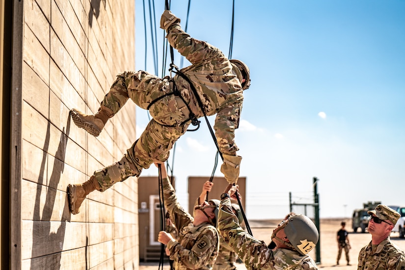 Soldiers help a fellow soldier rappel down a tower in a desert-like area.
