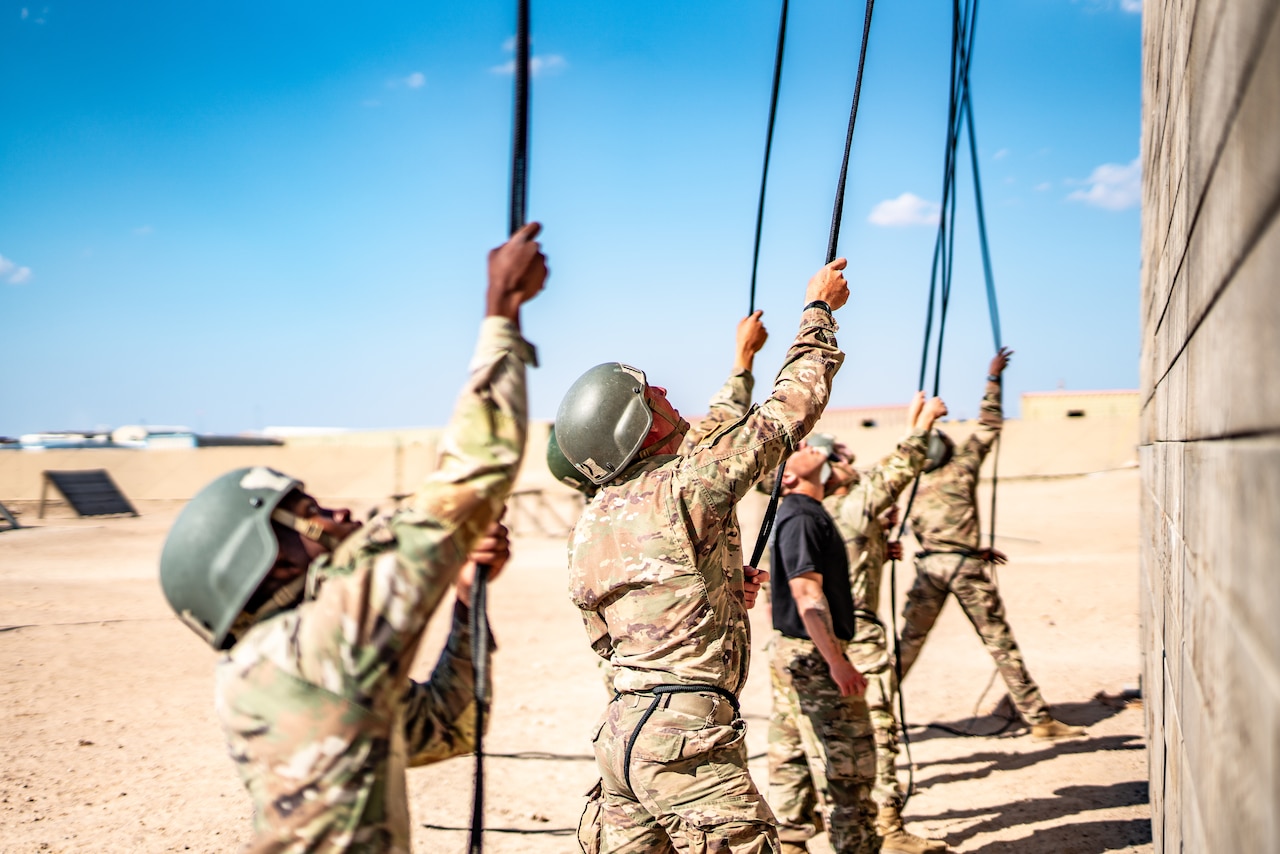Soldiers help fellow soldiers rappel off a tower in a desert-like area.