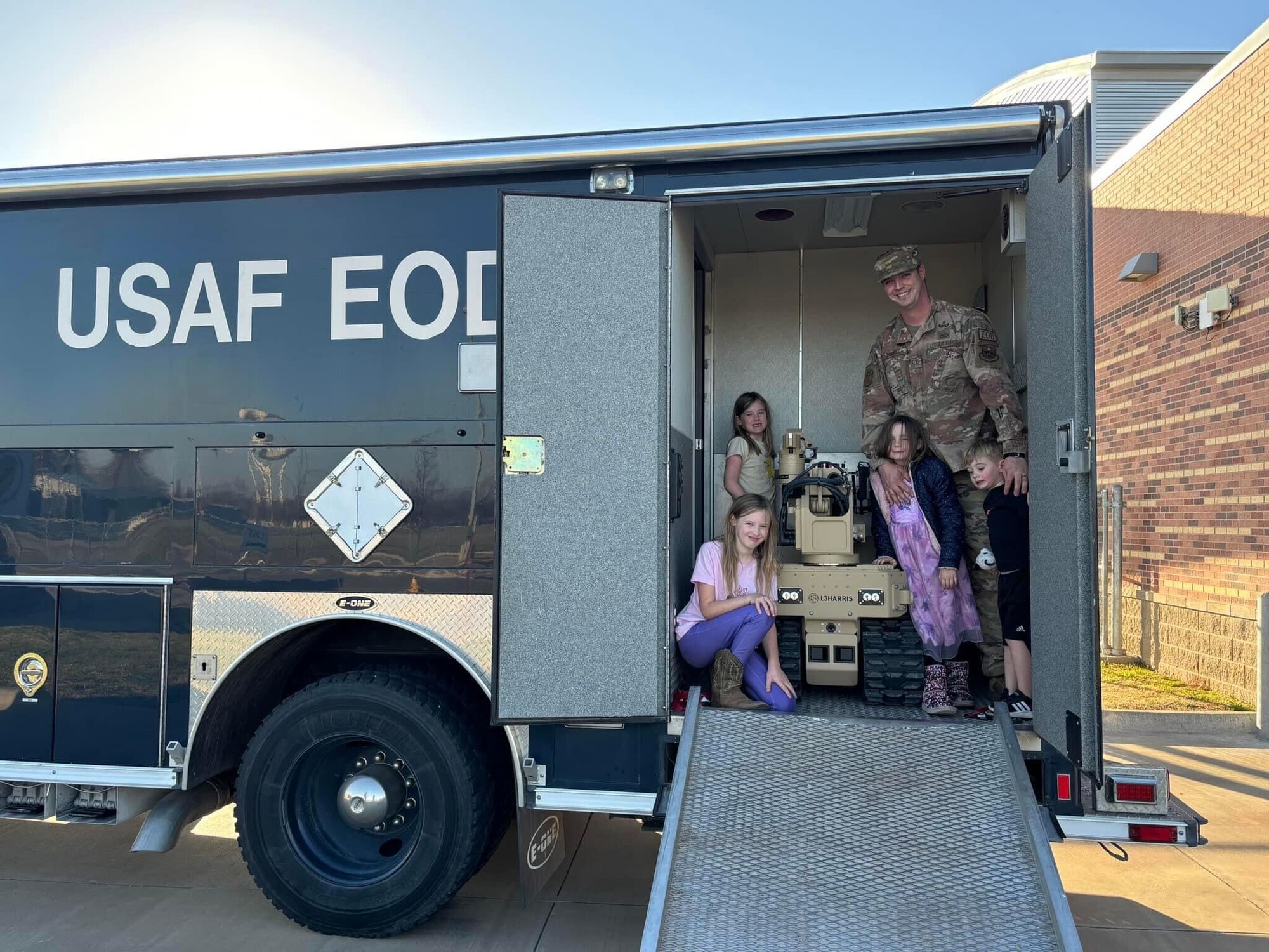 An Airman stands in a trailer with children and a robot.