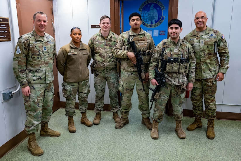 Service members stand in line for a photo with a commander in front of a blue bulletin board in an office hallway.