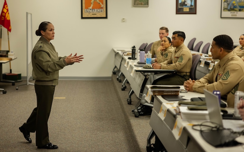 A Marine stands and gestures to seated Marines in a classroom setting.