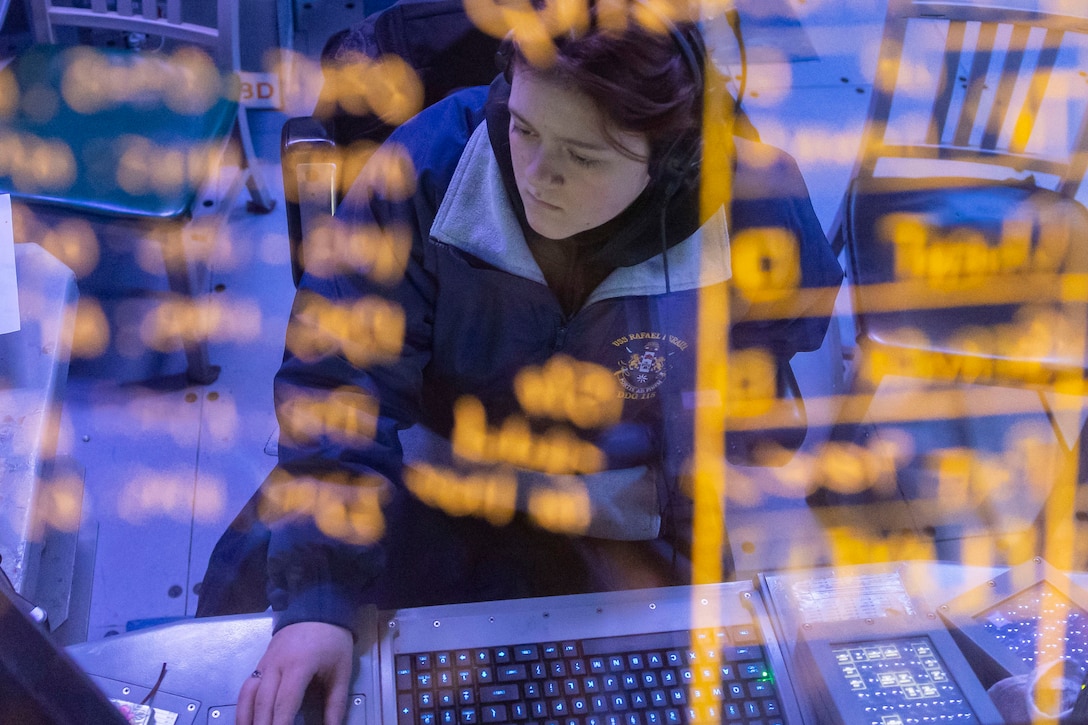 A sailor wearing a jacket is seen through a glass panel with yellow lettering using a computer and looking at a monitor.