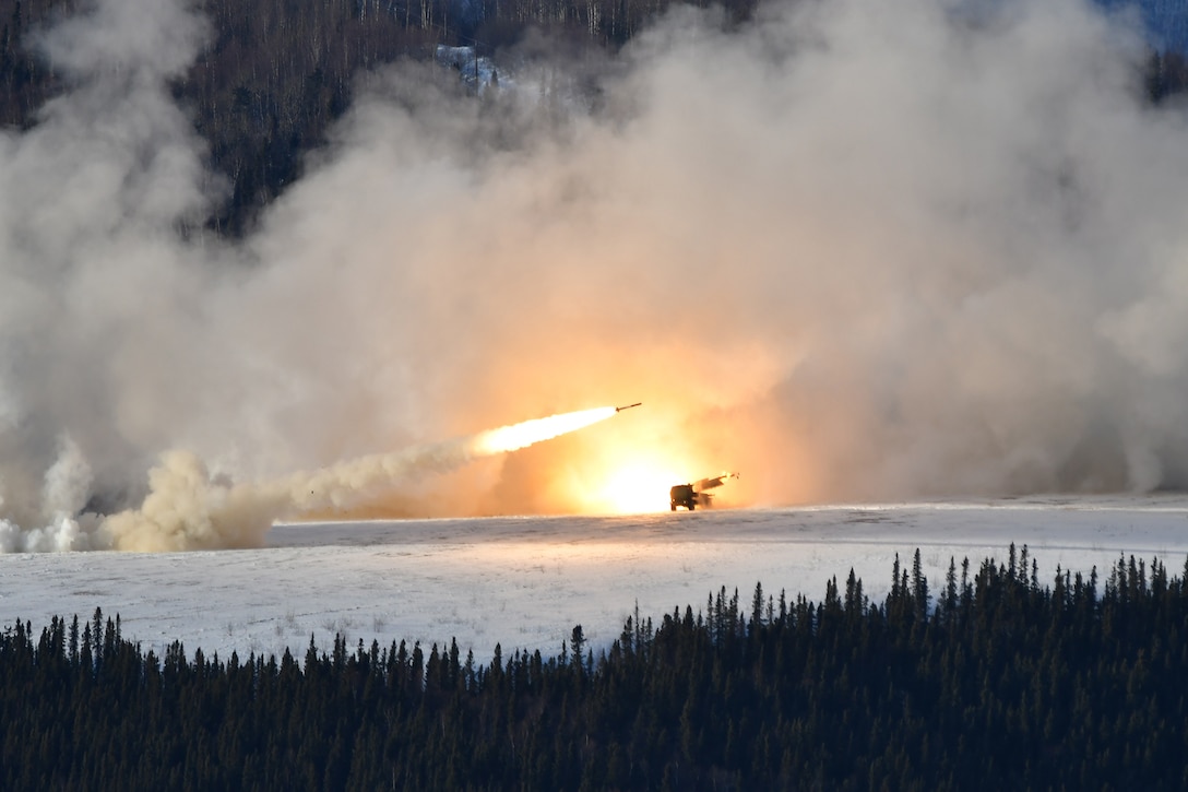 A rocket blasts from a high mobility artillery rocket system creating a large plume or orange and white smoke over snowy ground.