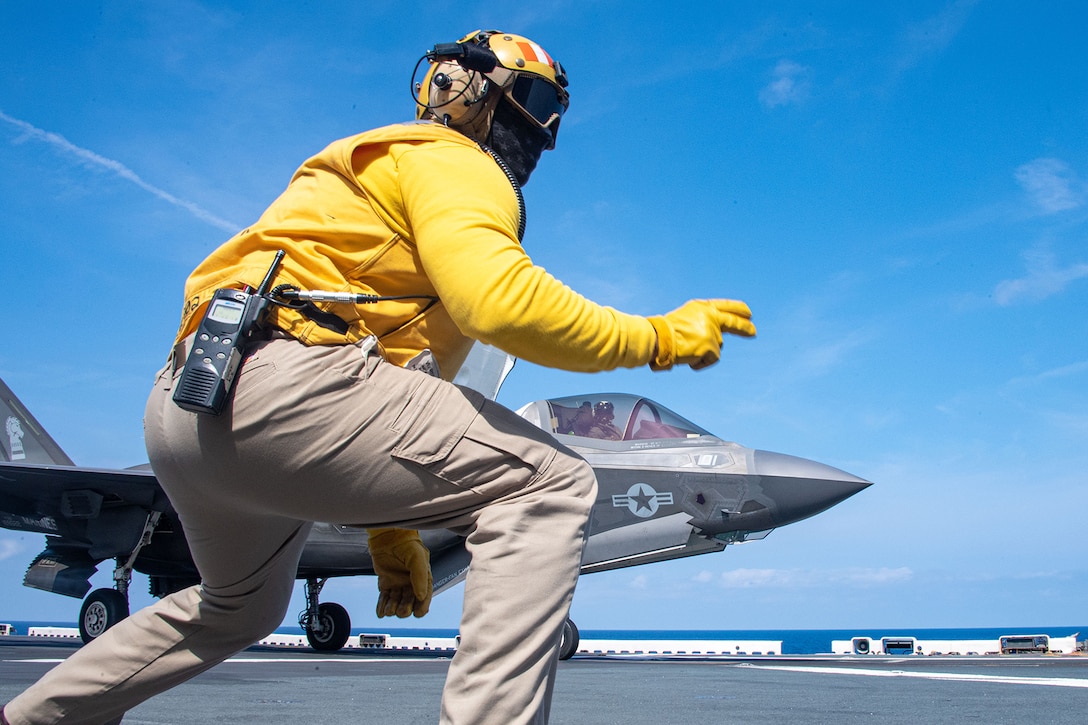 A sailor squats to signal a military aircraft as it prepares to take off from a Navy ship’s flight deck.