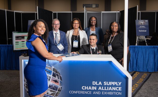 Six people pose for photo at blue and white booth. Woman in front left wearing blue dress.