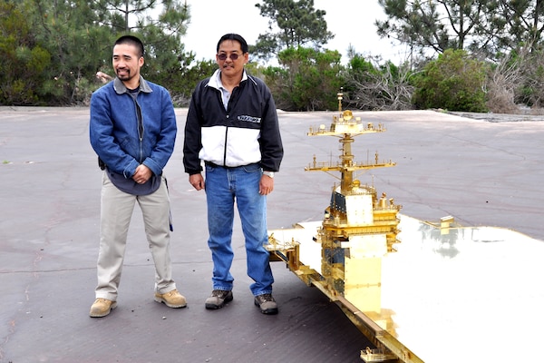 two men pose outside next to a partial view of a, intriciate, gold-colored brass model ship