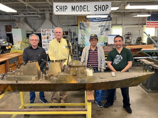 four men pose inside a ship model shop behind a brass ship model with intricate parts; a sign above them says “SHIP MODEL SHOP”