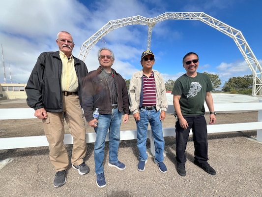 four men pose outside in front of a large white arch on a sunny day