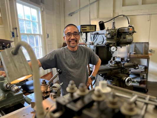 a man wearing glasses poses, smiling, in a workshop surrounded by tools and equipment