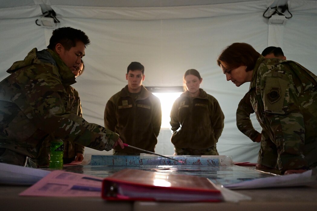 Inside a tent, a group of military members are pointing to and looking at key points on a map.
