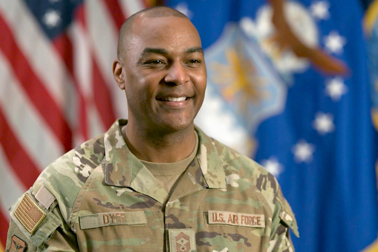 A person in uniform smiles during an interview in front of flags.