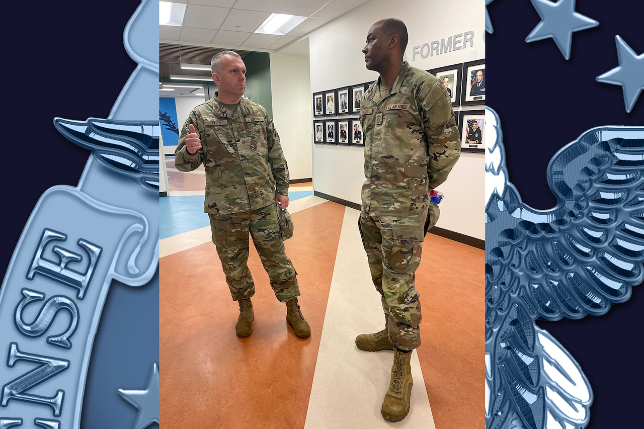 Two service members in uniform talk in a hallway in front of a wall of framed portraits.