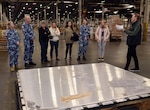 Photo is of a group of people, some in military uniform, standing around a large metal pallet in a large warehouse.