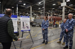 Photo is of a man and a group of people in military uniforms  standing in a warehouse in front of placards on easels.