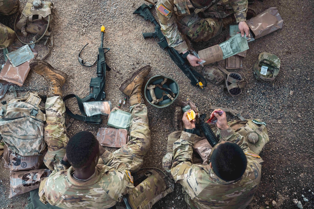 Soldiers sit on a dirt road surrounded by tactical gear while eating as seen from above.
