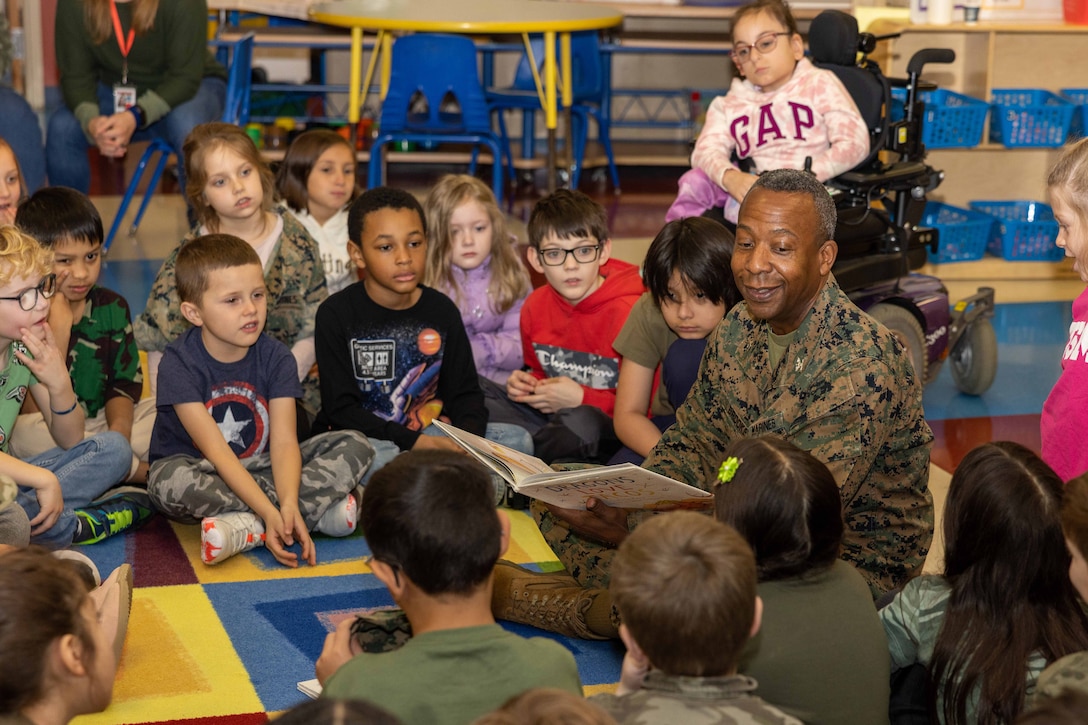A Marine Corps officer sits on a multicolored carpet and reads a book to a group of children.