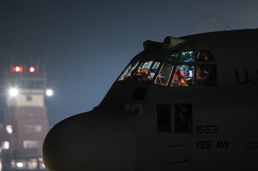 Airmen sit in the cockpit of a military aircraft shown in silhouette at night.