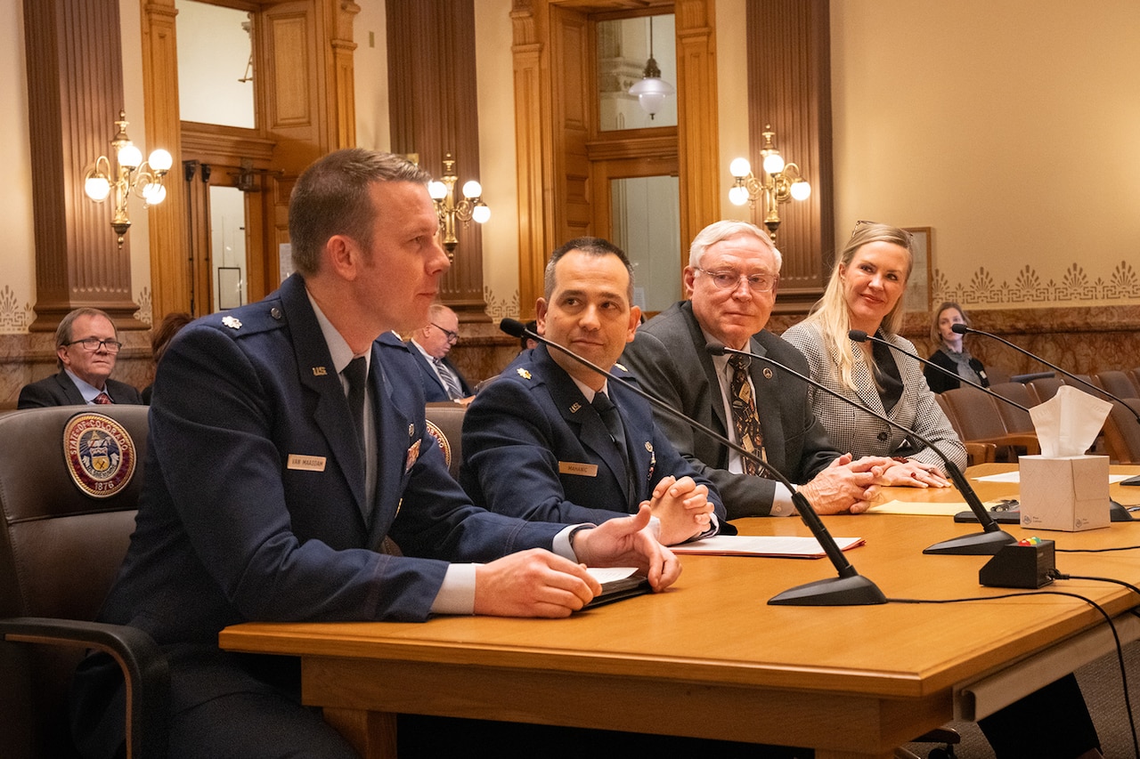 Two people in military uniforms are seated at a table with microphones next to two people in civilian business attire.