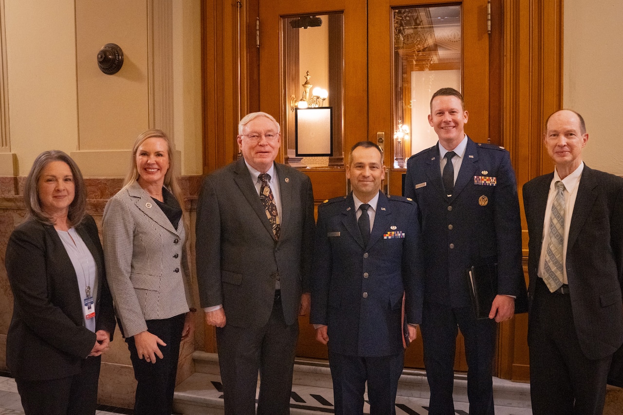 People in civilian business attire stand next to two service members in uniform.