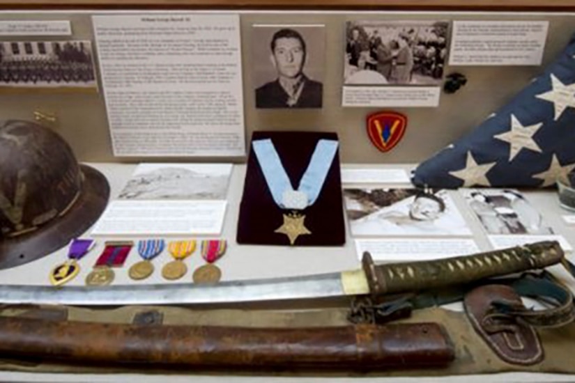 A display holds medals, a sword, a flag and a helmet of a person who is also pictured.