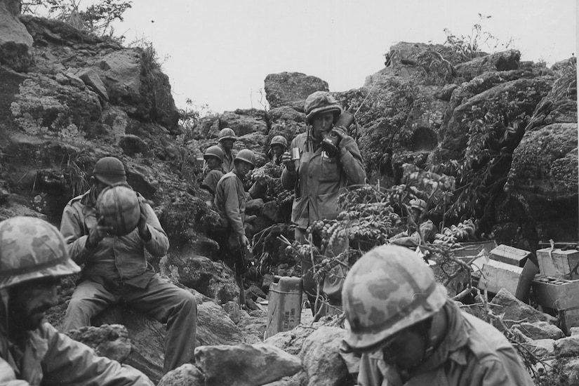 Service members sit and stand at ease in a rocky trench.