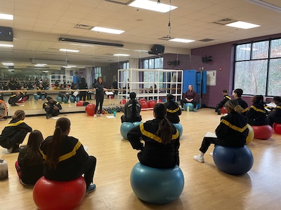 Women in military physical training uniforms sitting on rubber exercise balls in a gym listening to instructor.