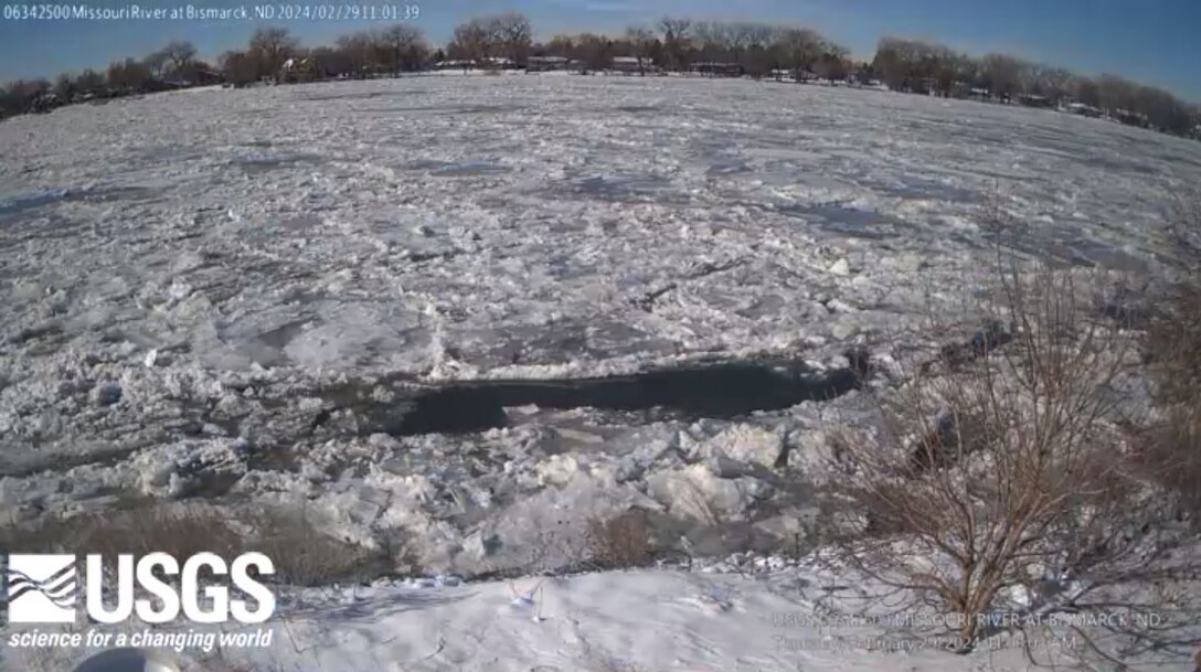 A fisheye lens view looking across the Missouri River in Bismarck, N.D. The foreground of the photo shows a snowy riverbank and some brush. The river is covered in thin ice that has buckled in some areas. Across the river is a treeline under blue skies.