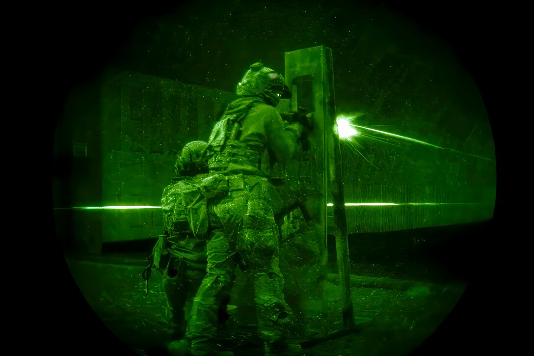 Two soldiers wearing tactical gear fire weapons while standing next to each other as seen through night vision lens.