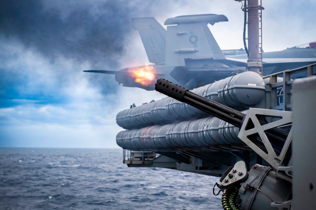 Flames and smoke shoot from a mounted weapon system fired from the side of an aircraft carrier at sea.