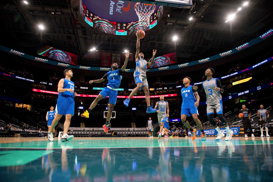 An airman shoots a layup during a basketball game in an arena as one defender tries to block it and other players watch.