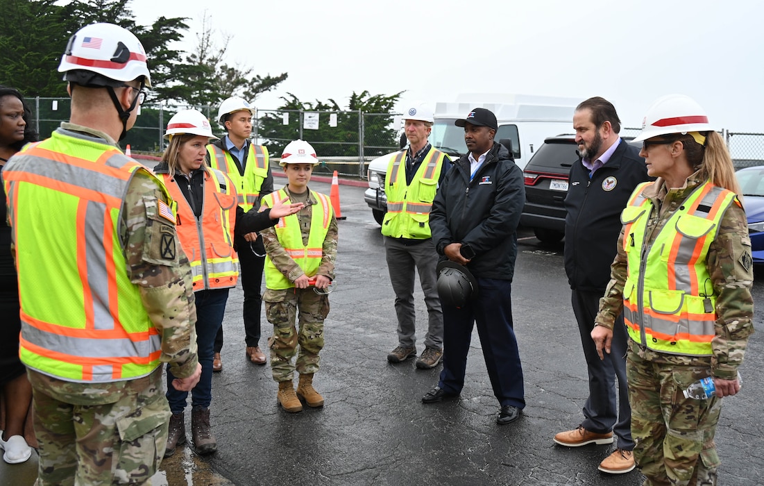 A group of people in orange safety vests and hardhats with two men in black jackets stand outside in a parking lot listening to a white female talking.
