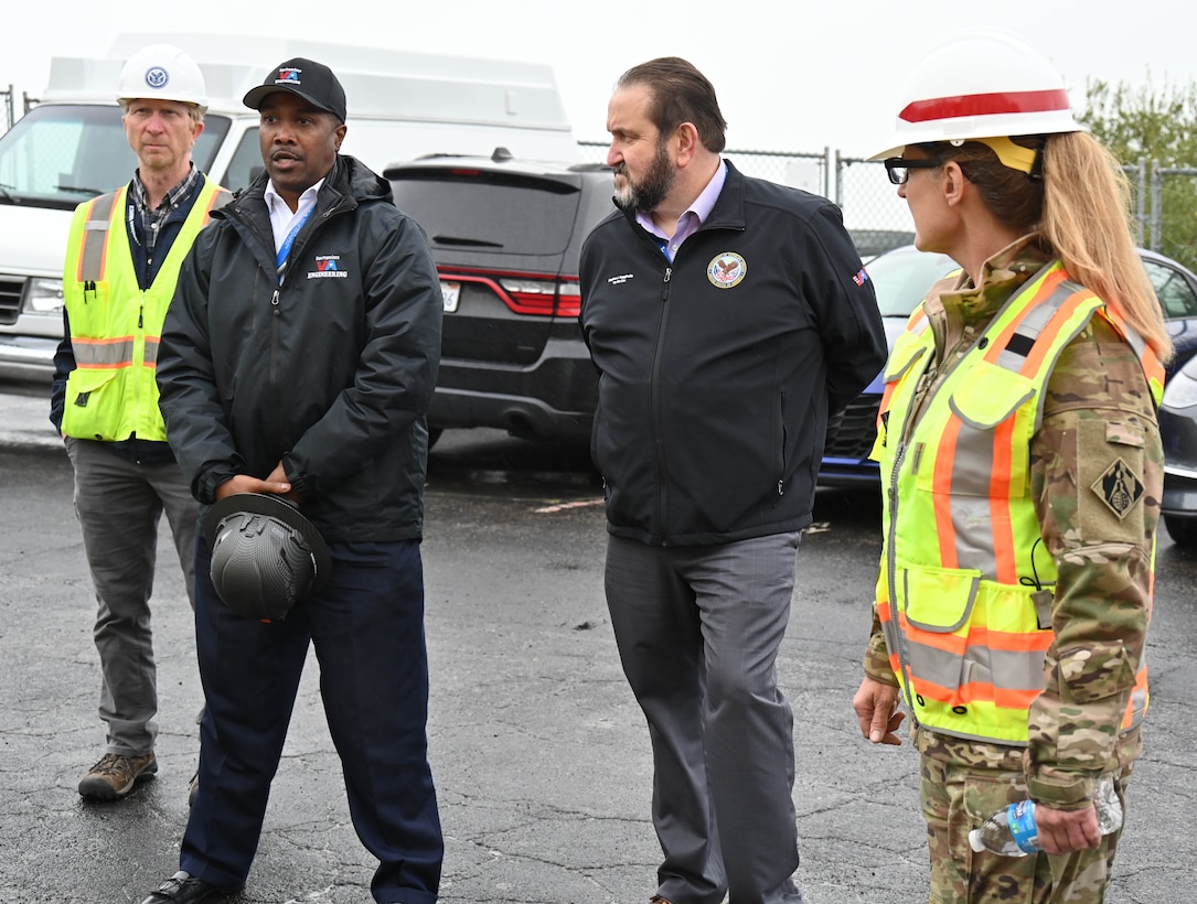 A black man in a black jacket stands next to a white man in a black jacket. They are both between a white man and a white female in orange safety vests and hardhats.