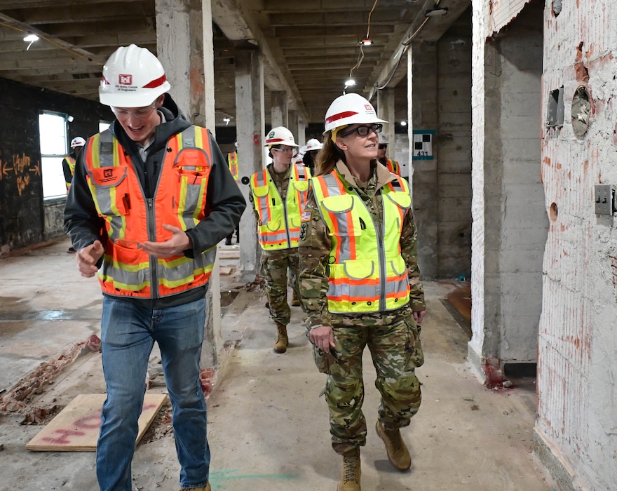 Three white people walk through a building construction site in orange safety vests and hardhats.